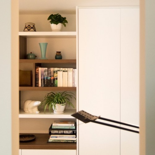 In een doorgang tussen de inkomhal en de living wouden de eigenaars van dit appartement een bureau inrichten. Tegenover het bureaublad plaatsten we een kamerhoge  kast waarin de technische ruimte met allerlei apparatuur en leidingen van het hele appartement weggestopt zit. Om de indruk te wekken dat deze ruimte enkel als bureau dient, creëerden we naast deze hoge kast nog een lage kast. Om het geheel een speelse look te geven combineerden we enkele bijpassende zwevende leggers in wit en Notelaar. Enkele verticale én verplaatsbare tussenschotten zorgen ervoor dat de bewoners kunnen variëren met hun decoratie. Kast-ID Asse